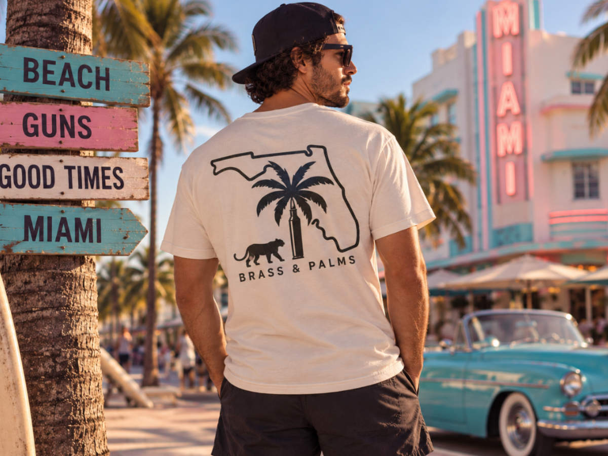 A man wearing a beige graphic t-shirt with a black Florida state outline, a panther and palm tree-bullet design, standing on a sunny beach boardwalk with vintage cars and palm trees in the background.