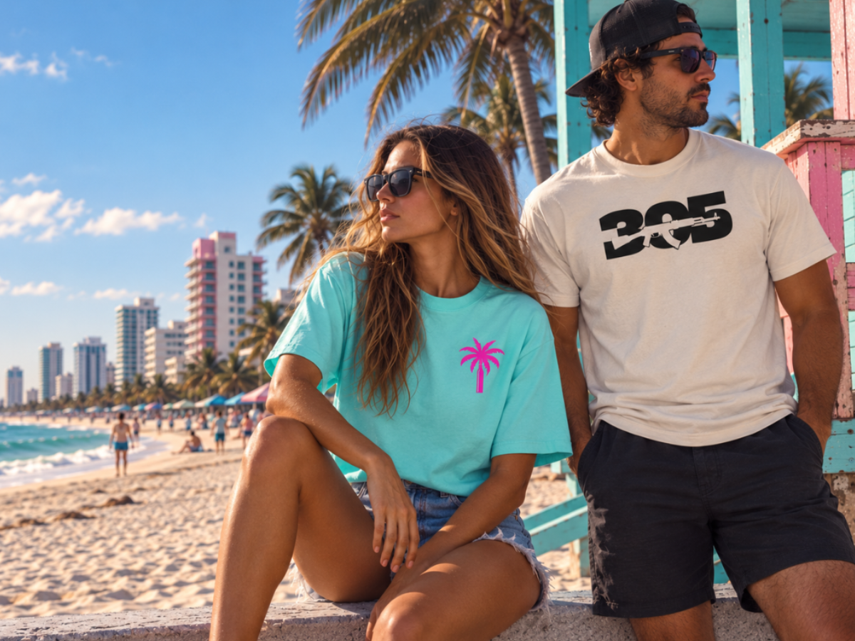 A man and a woman sitting on a beach wall, wearing matching graphic t-shirts. The woman wears a light turquoise t-shirt with a pink palm tree graphic, and the man wears a cream t-shirt with a black '305' with rifle design graphic.