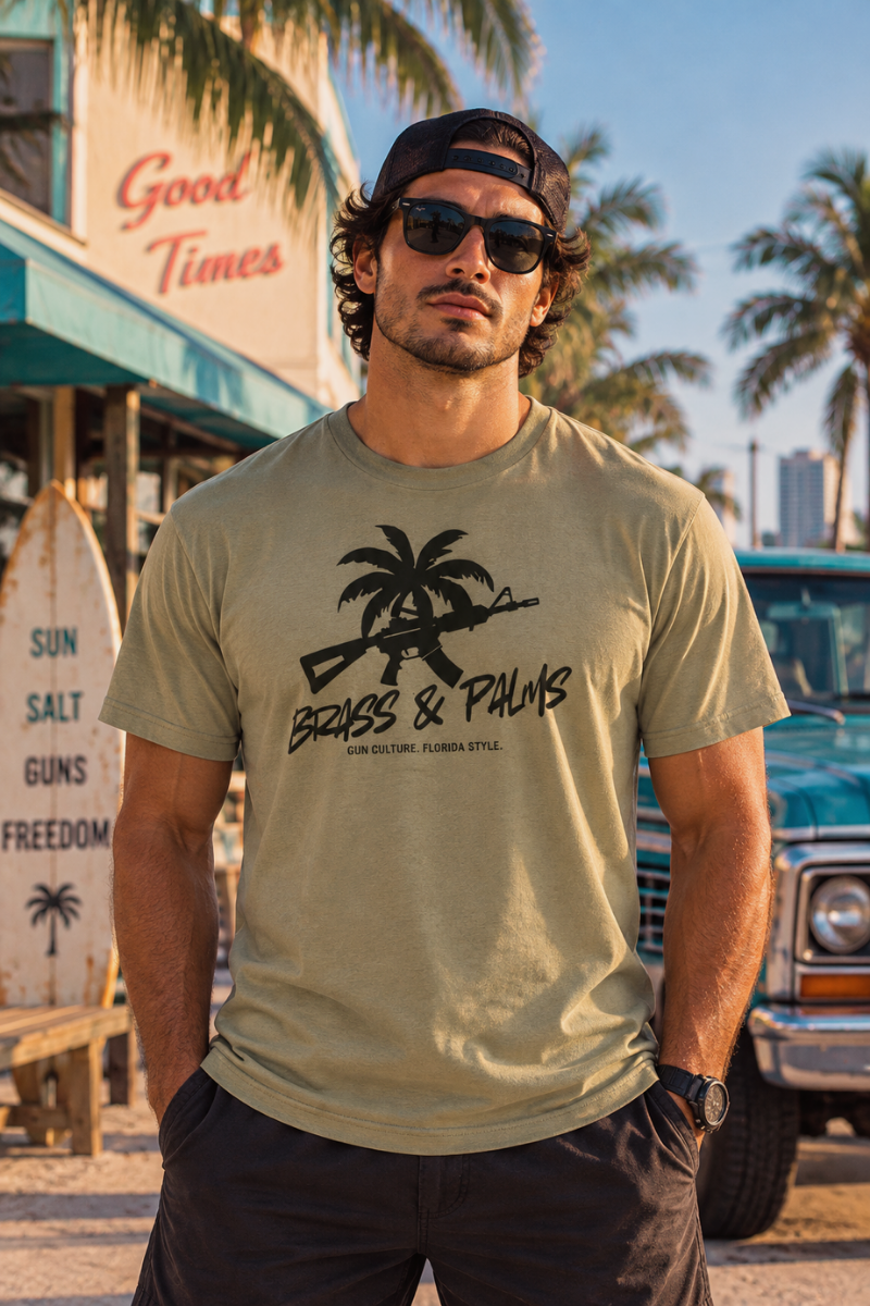 A man wearing a beige graphic t-shirt with a black palm tree and rifle design, standing outdoors in front of a tropical-themed building and a vintage blue truck.